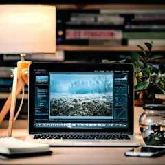 Laptop z edytorem zdjęć, na ekranie zimowy krajobraz. Obok lampka, notatnik i słoik z przetworami. To idealne miejsce do pracy nad plikami tif.