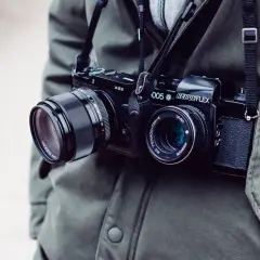 Osoba w zielonej kurtce nosi na szyi dwa aparaty fotograficzne: jeden cyfrowy, drugi analogowy.