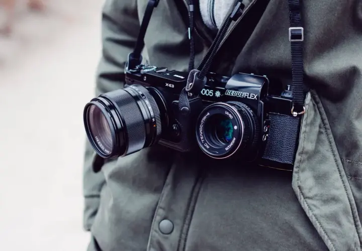 Osoba w zielonej kurtce nosi na szyi dwa aparaty fotograficzne: jeden cyfrowy, drugi analogowy.