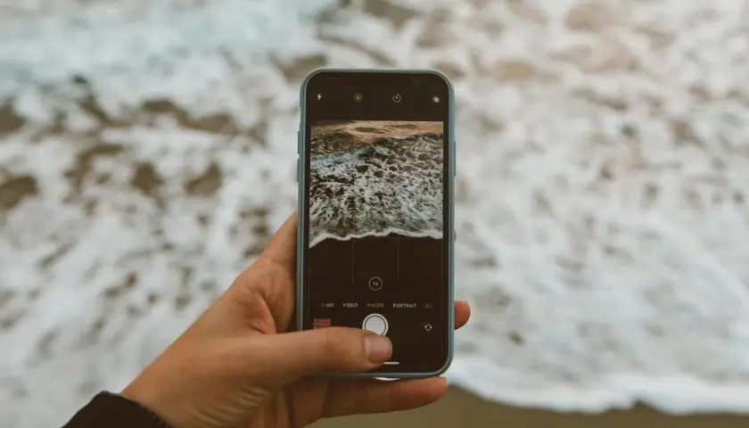 Dłoń trzyma smartfon, którym fotografuje fale. **Expert raw** uchwyci piękno natury.